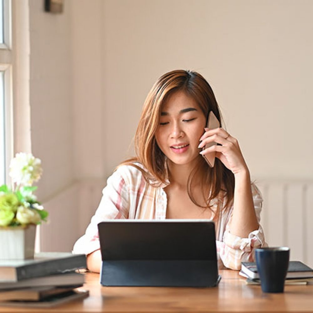 Photo a beautiful woman using a smartphone to calling someone while sitting in front a computer tablet with keyboard case at wooden working table over comfortable living room as background.